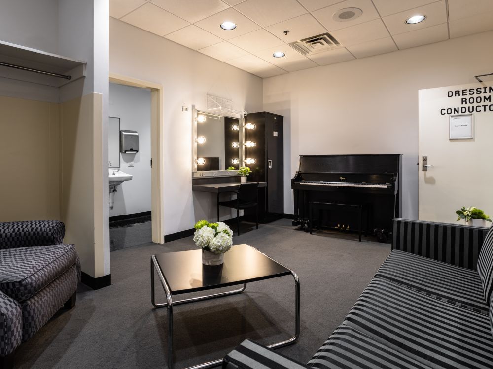 Image of Dressing Room 1 filled with a piano, a vanity mirror setup, a couch, accent chair, and small table fill the room. A sink indicating a bathroom is shown.