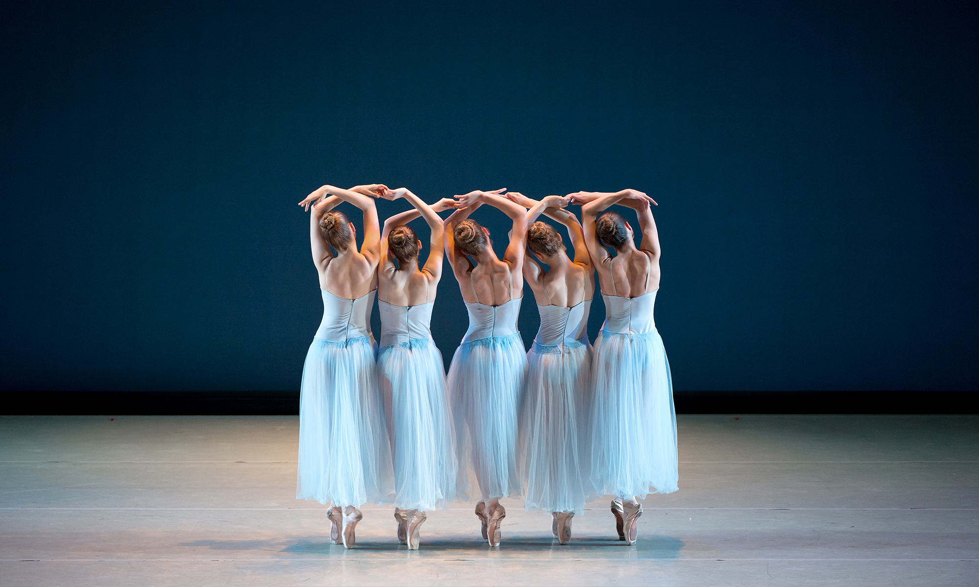 Five dancers wearing light blue tulle dresses raising their arms up