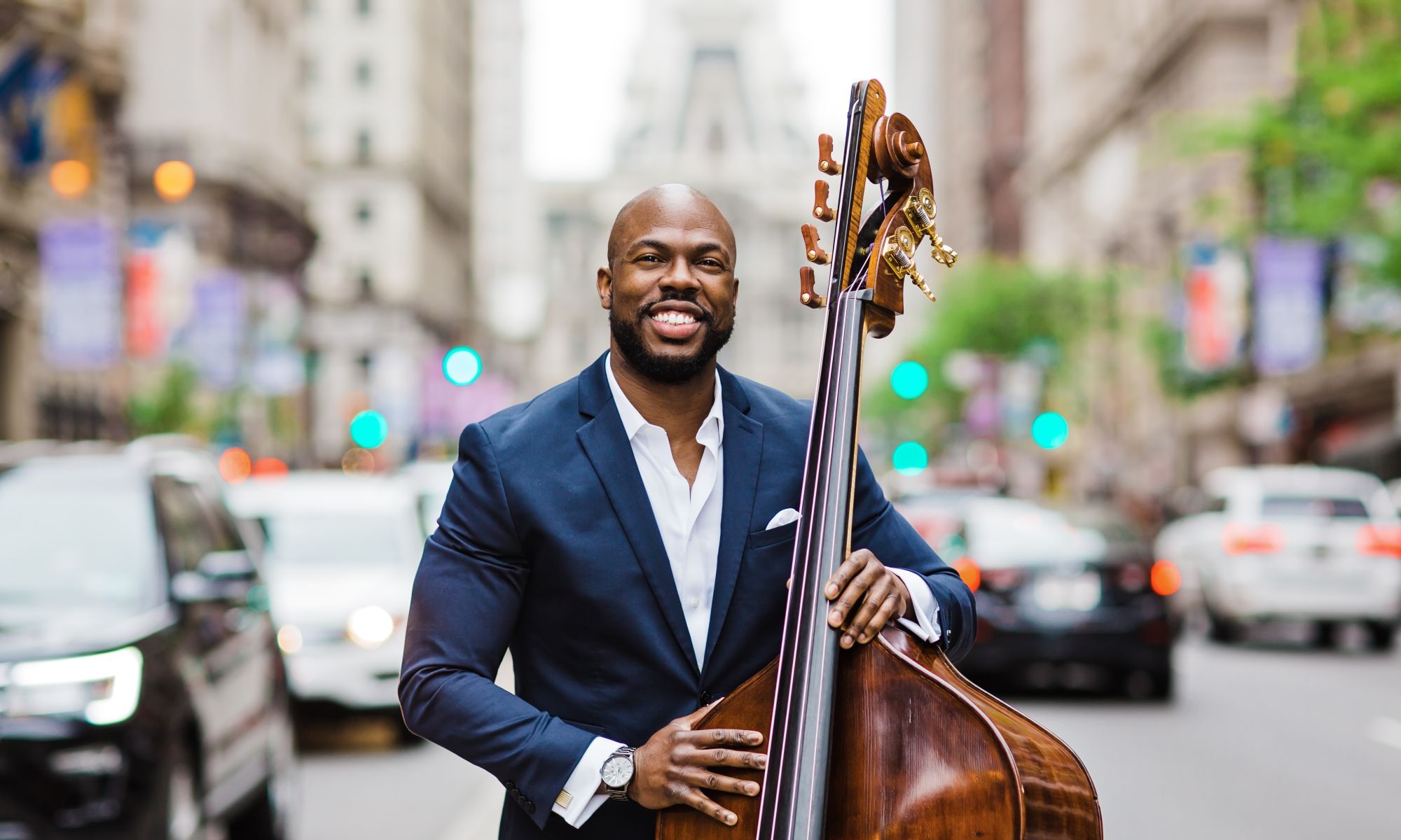 Joseph Conyers wears a blue suit with a white shirt, holding a string instrument in a busy city street