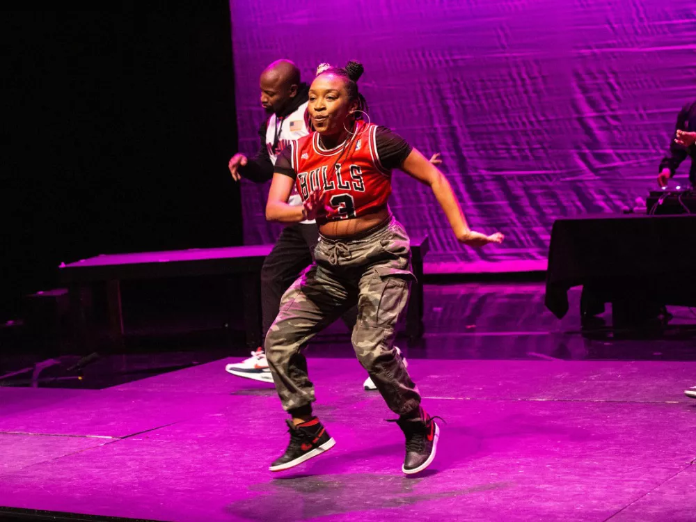 Image of Donetta “Lilbit” Jackson dancing on a brightly colored stage. Donetta appears mid movement with her hands shuffling from the front to the side of her body. She wears a Chicago Bulls’ jersey, a black undershirt, and camo-colored pants. The stage is lit a bright magenta, deepening the black shadows in the background. Other dancers are in the background and a dj attentively looks at his turntables.