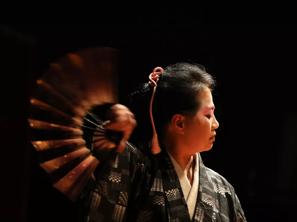 Image of Rika Lin and fan in motion. Rika’s profile faces the right side of the image, and she appears pensive. The other side of the image includes the moving brown fan which appears in contrast with the stark black background.