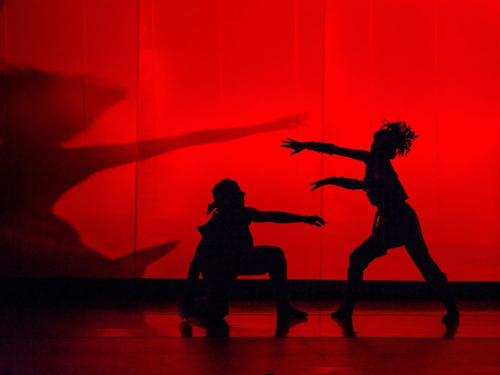 Two silhouettes of dancers performing in front of a red wall.  