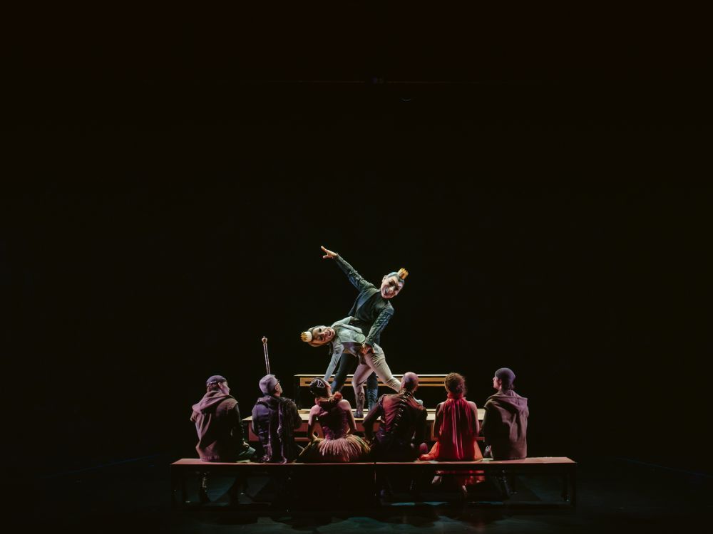 Two dancers on top of a table wearing puppet masks while six other performs watch them dance.