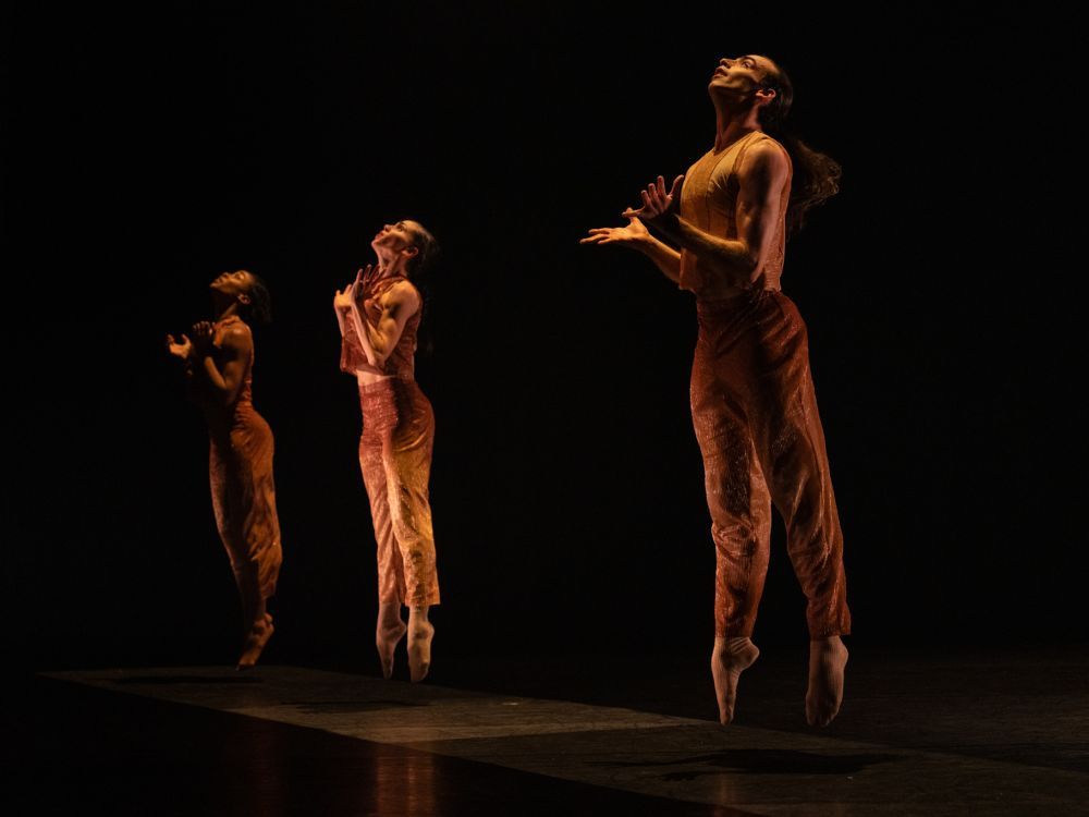 Three dancers on stage jump in unison while looking up.