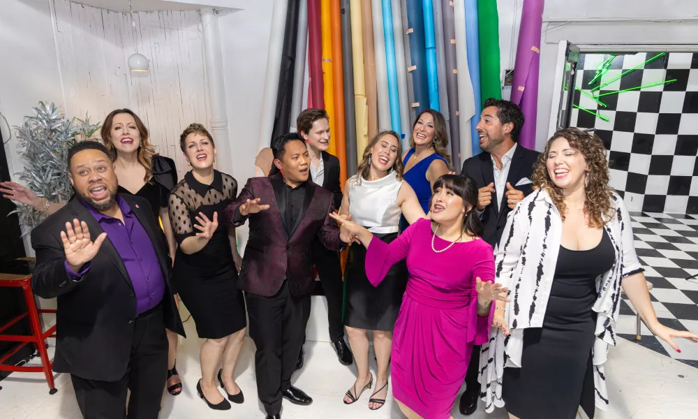 Group of ten singers singing and dancing in a room with fabric reams and checkered paint in the background.