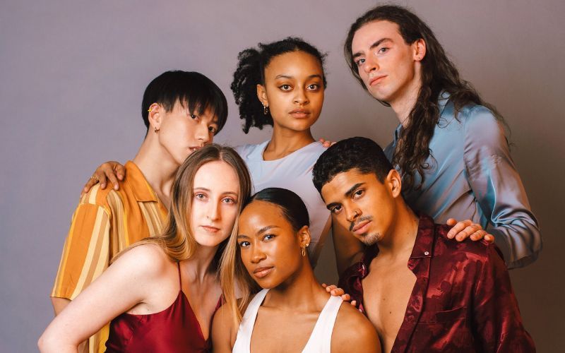 A group of dances pose together in front of a solid purple grey backdrop. The group wear color a range of autumn colors. 