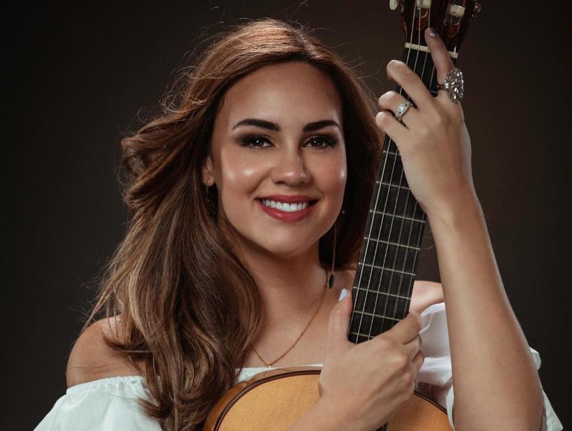 Portrait of Lisvette Sans holding a guitar in front of a dark brown backdrop 