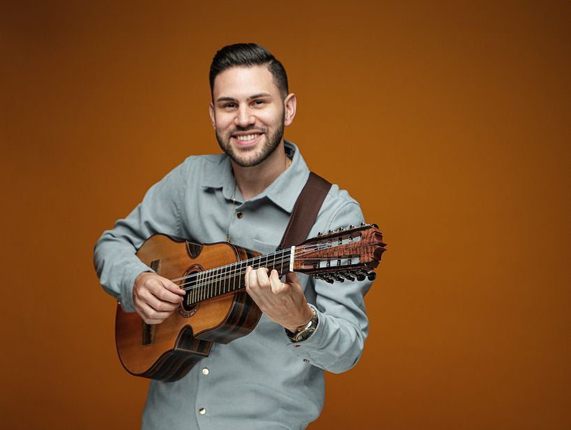 Portrait of Luis Sans holding a cuatro in front of a light brown backdrop. 