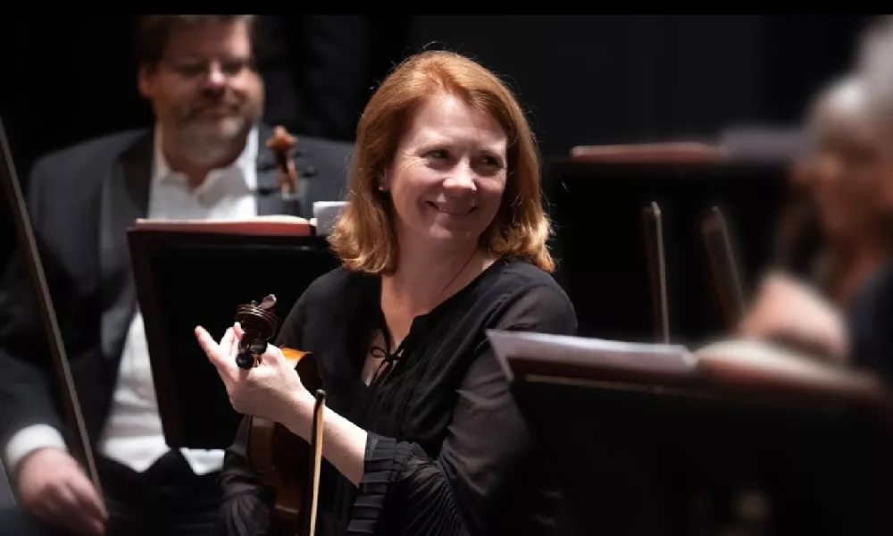 A woman with red hair smiles and holds a violin. 