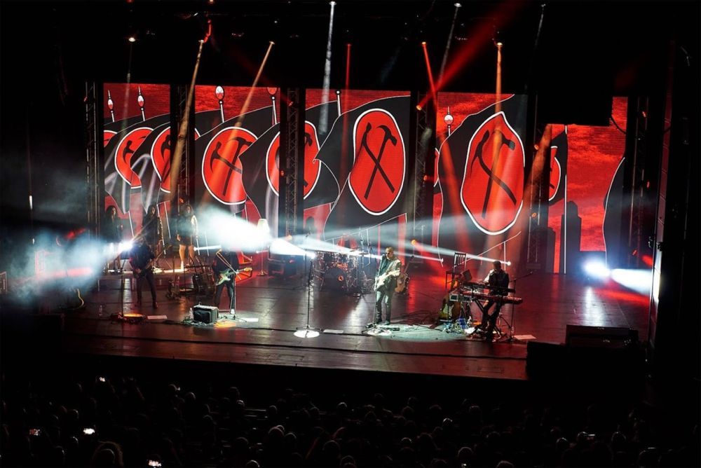 Rock musicians on stage. A flag with crosses axes appears on the large screens behind them.