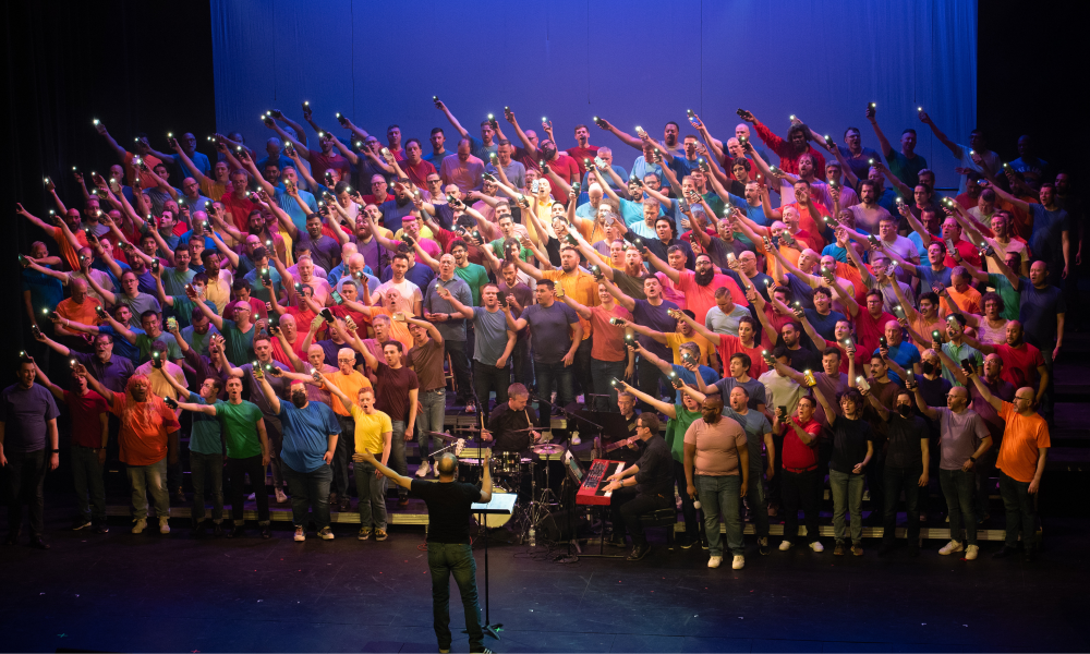 Over 200 CGMC members stand onstage on risers holding lights in their right hands. All hands are pointing upward and to the left. All members are wearing multicolored, bright t-shirts. The Artistic Director stands in front of the members wearing a black shirt and jeans. This is all set against a purple, lit background.