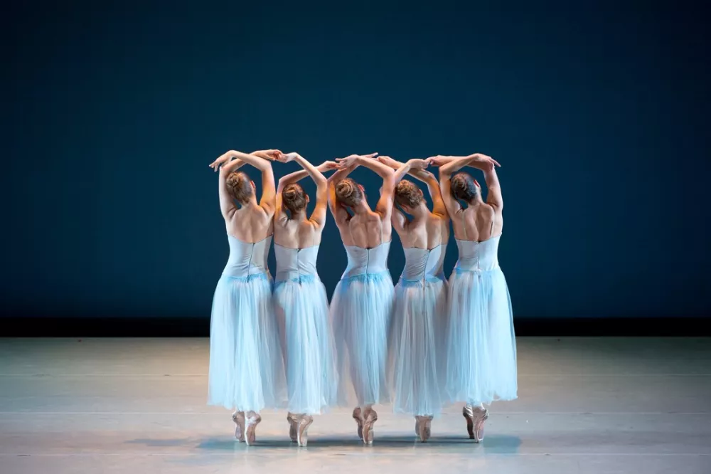 The back of five ballerinas in baby blue tulle tutu. They all create o's with their arms above their heads.