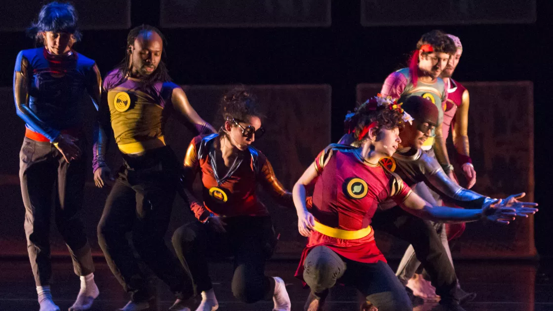 Seven Lucky Plush dancers in red, blue, purple, and yellow superhero outfits, 3 crouching and 4 standing with left arms reached out. 