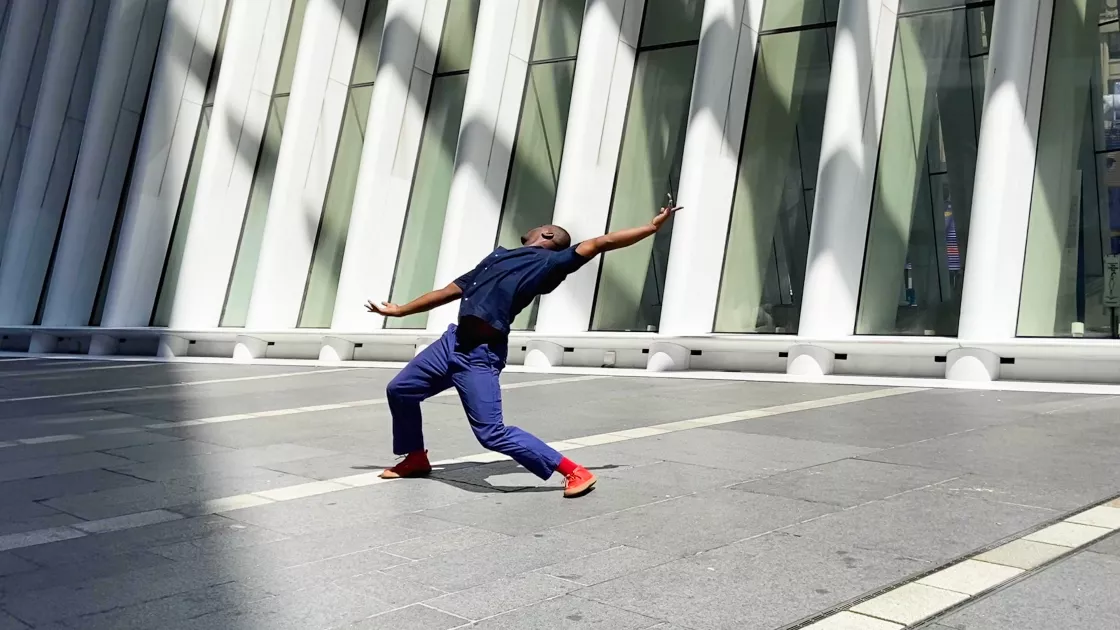 Dancer wearing blue jeans, blue button-up shirt, and orange tennis shoes stands in an empty, sun-filled parking lot. Dancer's legs are spread wide as he leans dramatically backwards, arms extended to his sides and head angled away from the camera. 