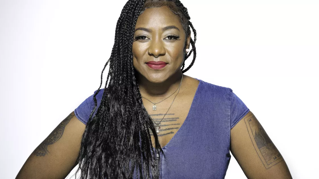 A Black woman with long braids and a blue shirt poses smiling, with her hands at her hips