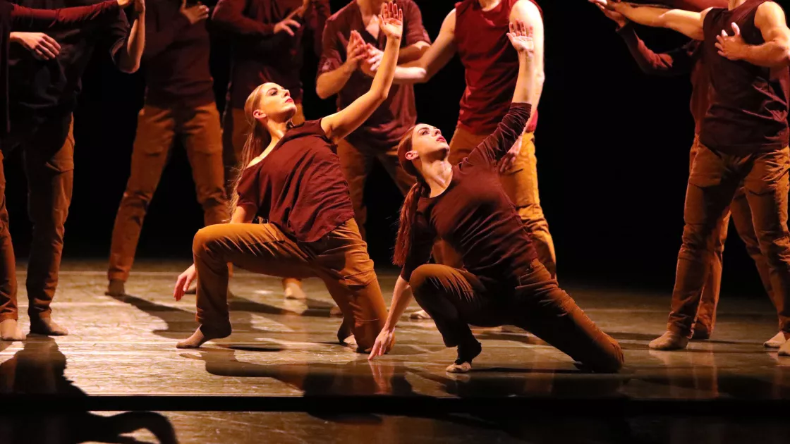 Dancers in brown sleeveless shirts and pants appear in standing and lunging positions on a black stage