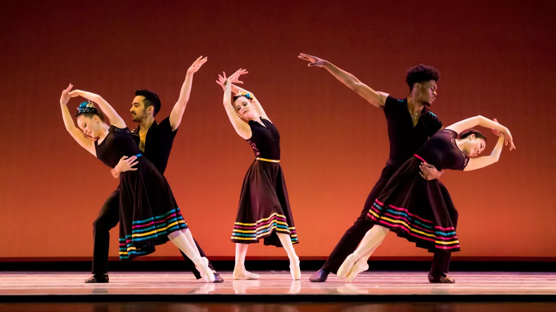 Two male and three female dancers dressed in black move against a burnt orange stage backdrop