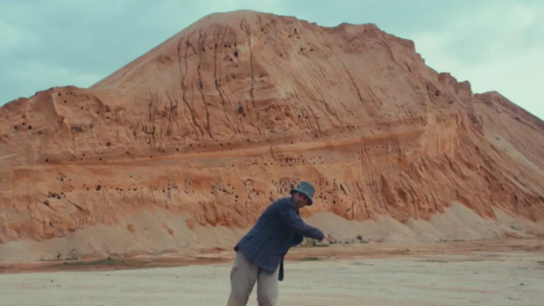 Dancer wears a blue bucket hat with a blue long-sleeved shirt and khaki pants, standing in front of a tan mountain