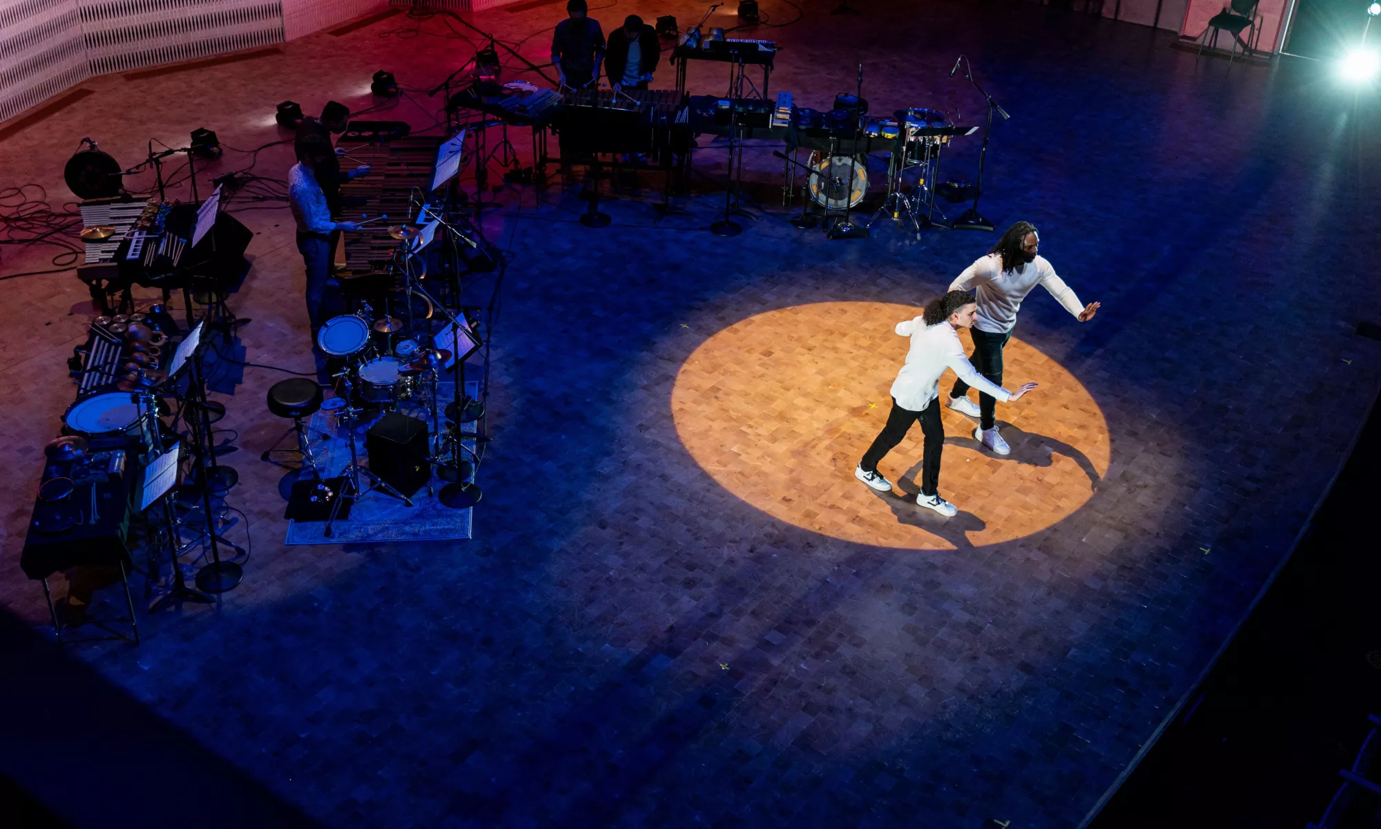 Two dancer dressed in white long sleeved shirts and black skinny jeans with white sneakers raise one arm forward, facing the audience