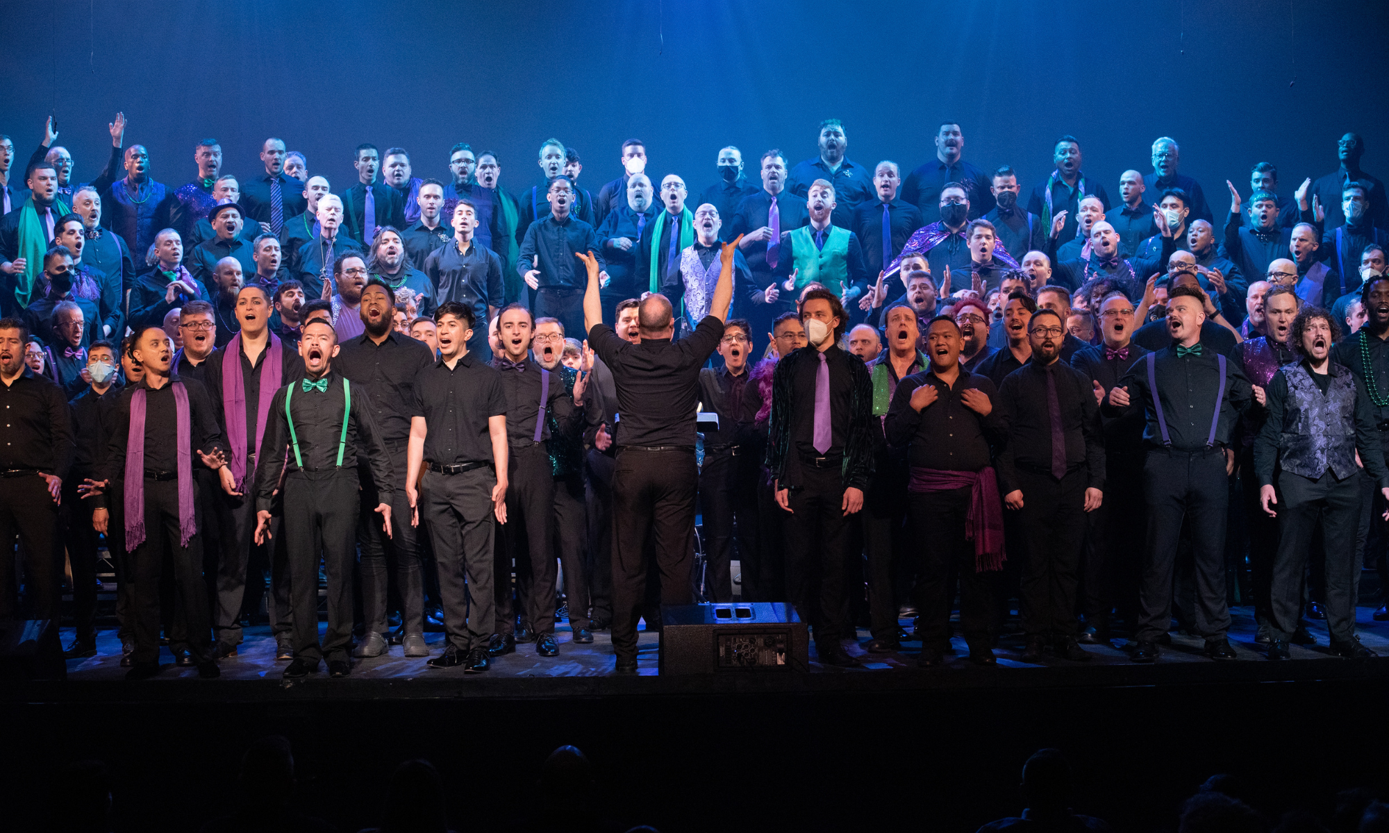 Picture of the chorus performing together onstage; members are dressed in all blacks with pops of green and purple color. Their mouths are open in song. The conductor stands at the front and center, back to the audience, arms stretched upward.