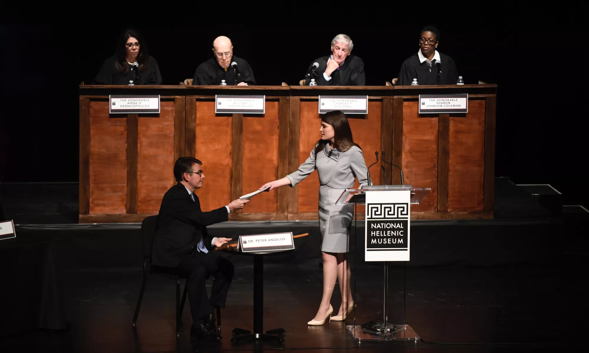 Image of participants on a stage, one is handing a file to a seated participant, with four judges in the background observing.