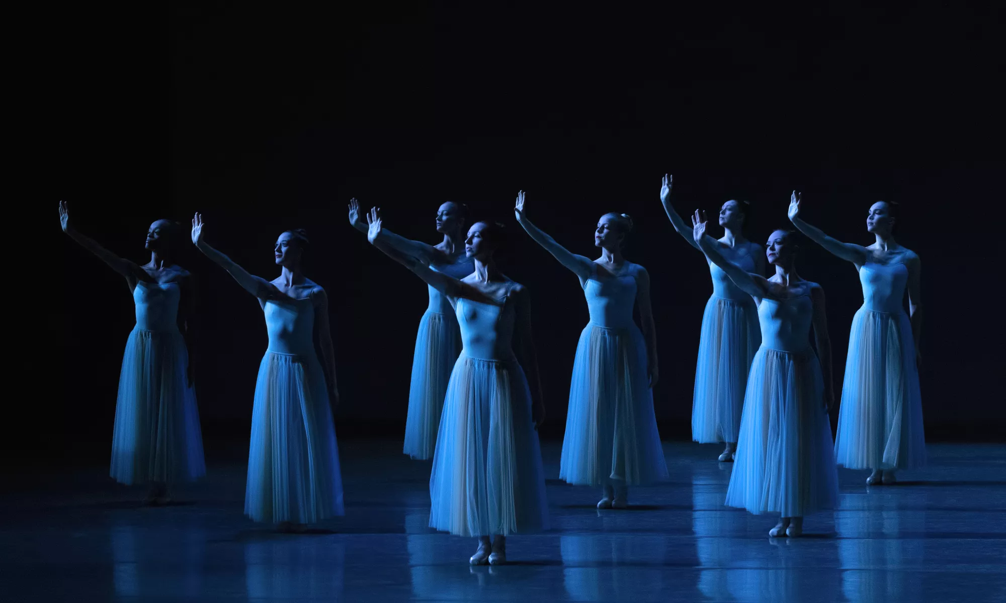 Several dancers dressed in white dresses lift their arms to stage right.