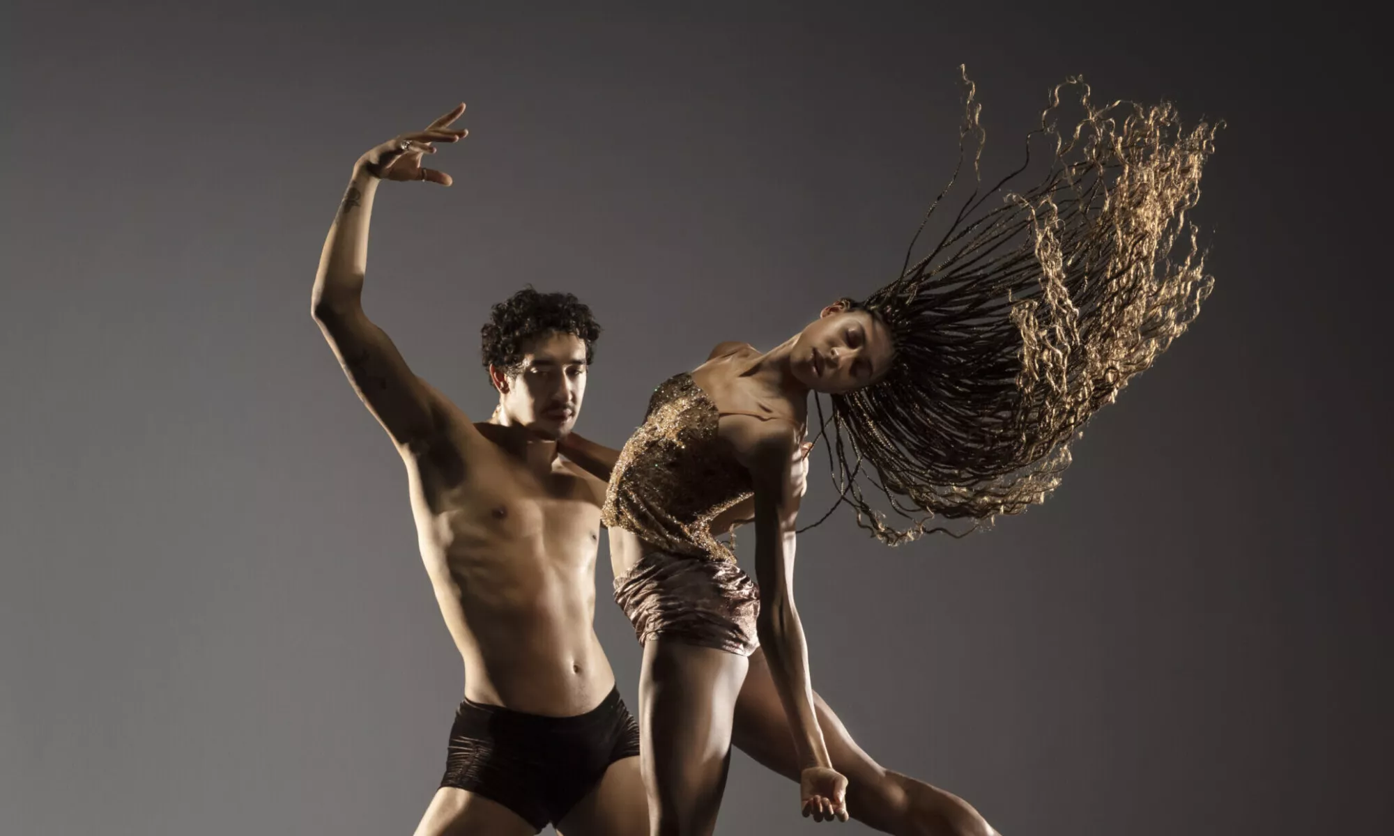 Male dancer wears black shorts, holding female dancer wearing a brown crop top and brown shorts