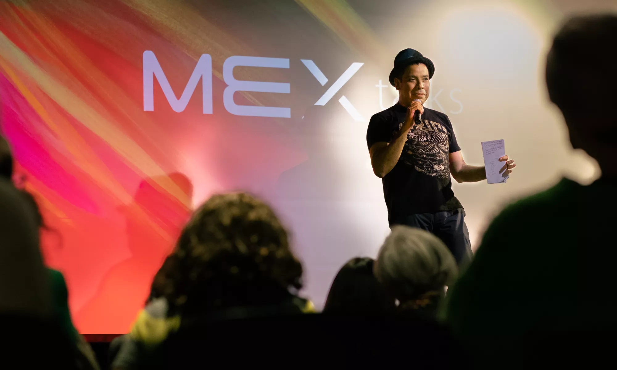 A man with a fedora hat stands in front of a crowd speaking into a microphone in front of a wall that projects the text "MEX talks"