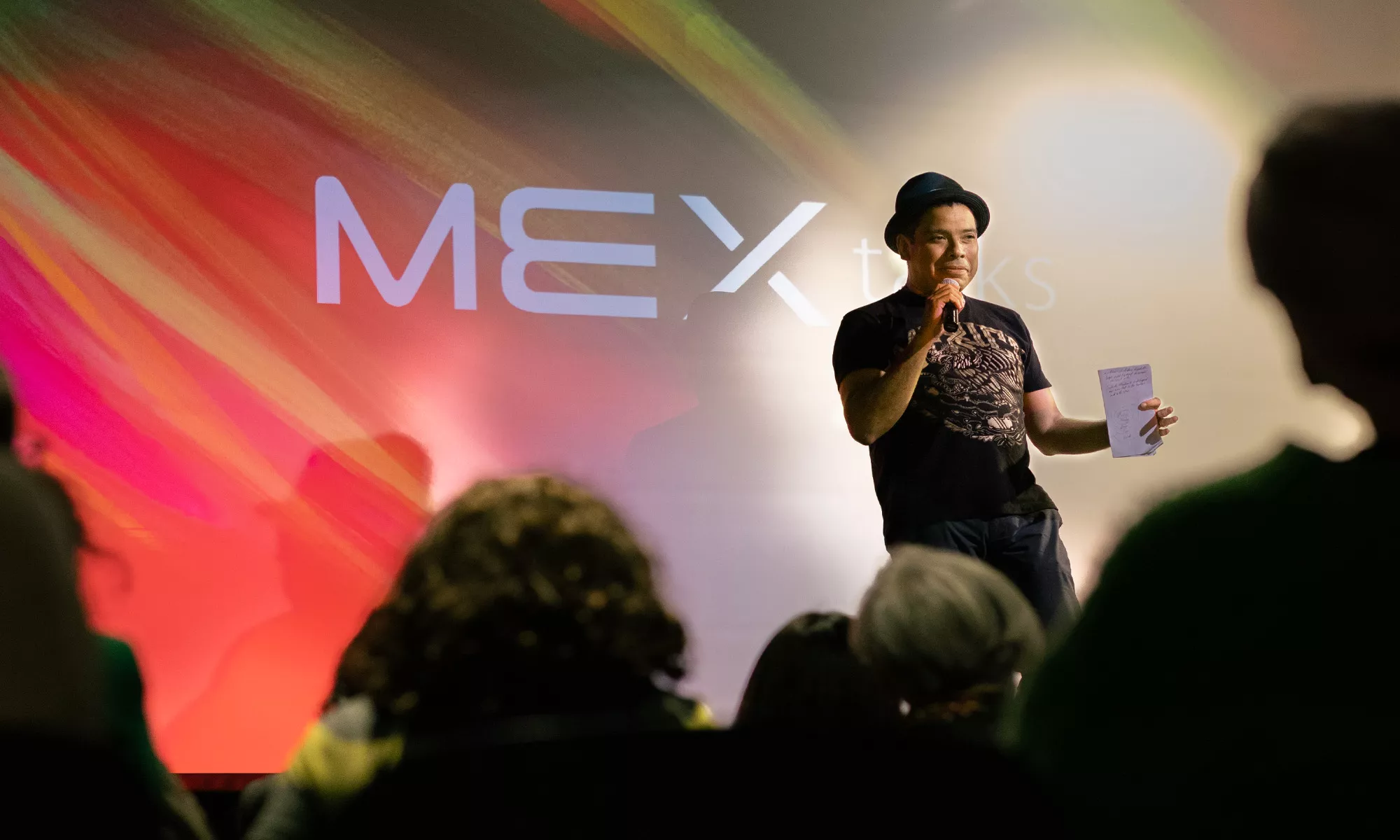 A man on stage holding a piece of paper speaks in front of crowd. The background is a multi-colored, primarily red image with the logo that reads "MEX talks".