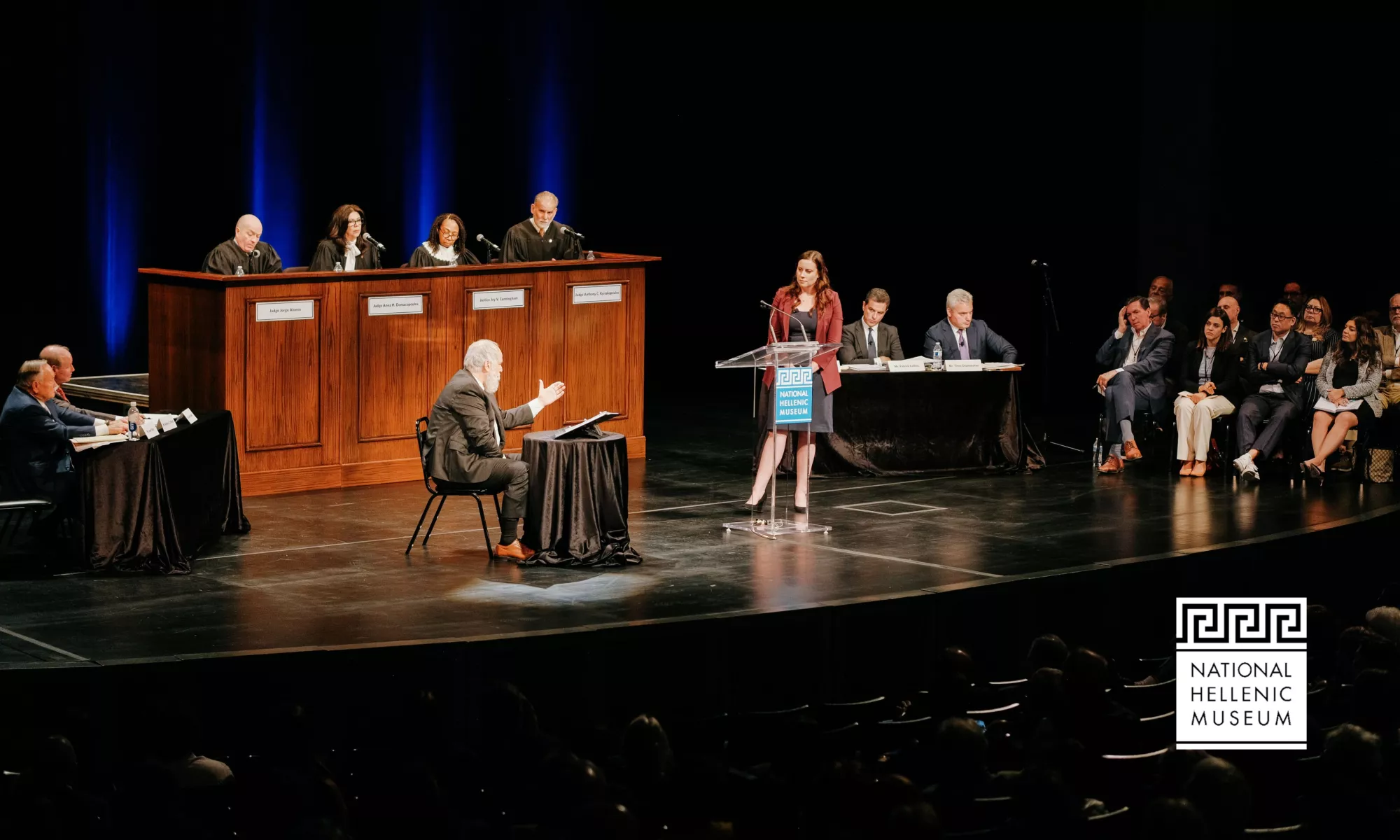 On a stage, a courtroom like setup takes place. 