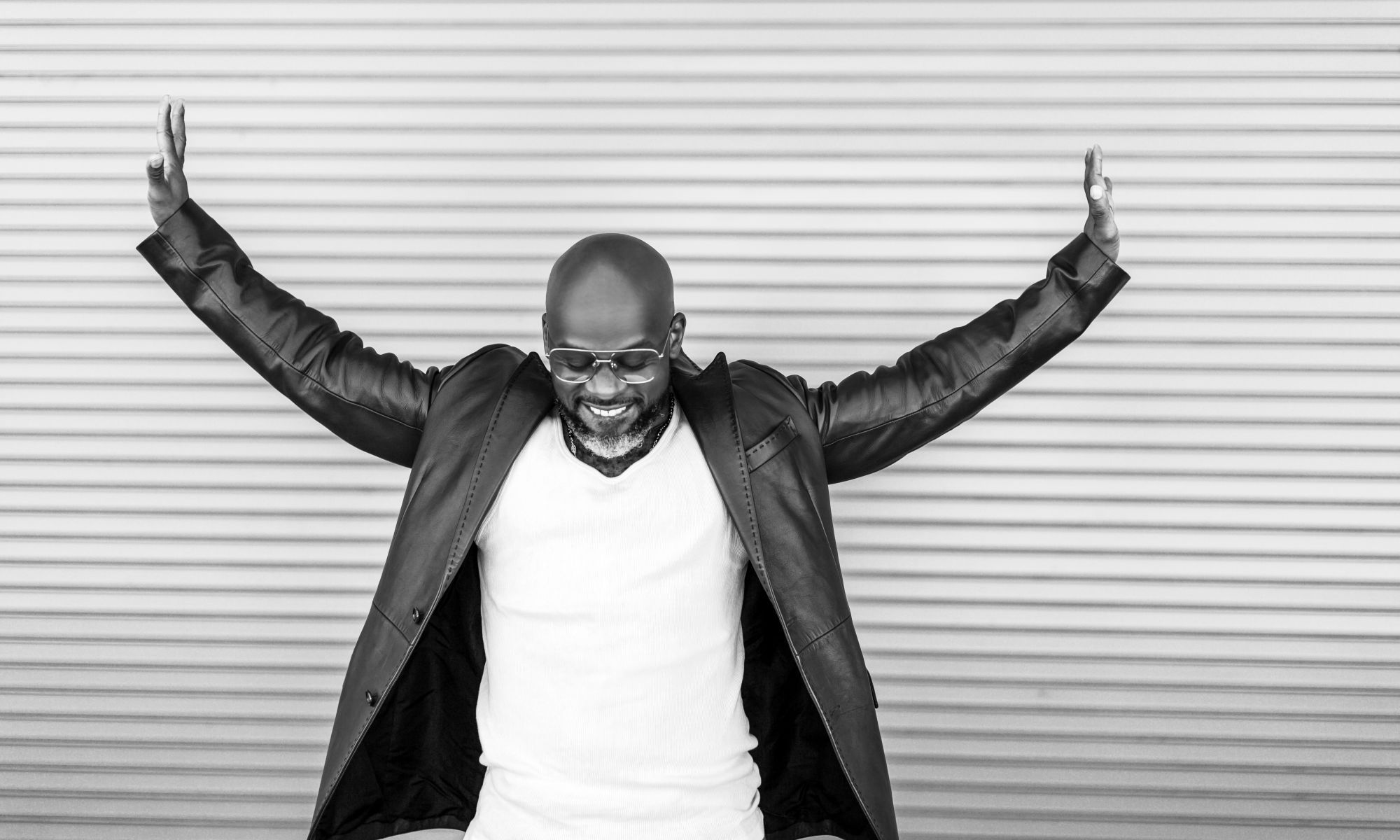 A black and white image of Marc Bamuthi Joseph while he lowers his face and extends his arms outward.