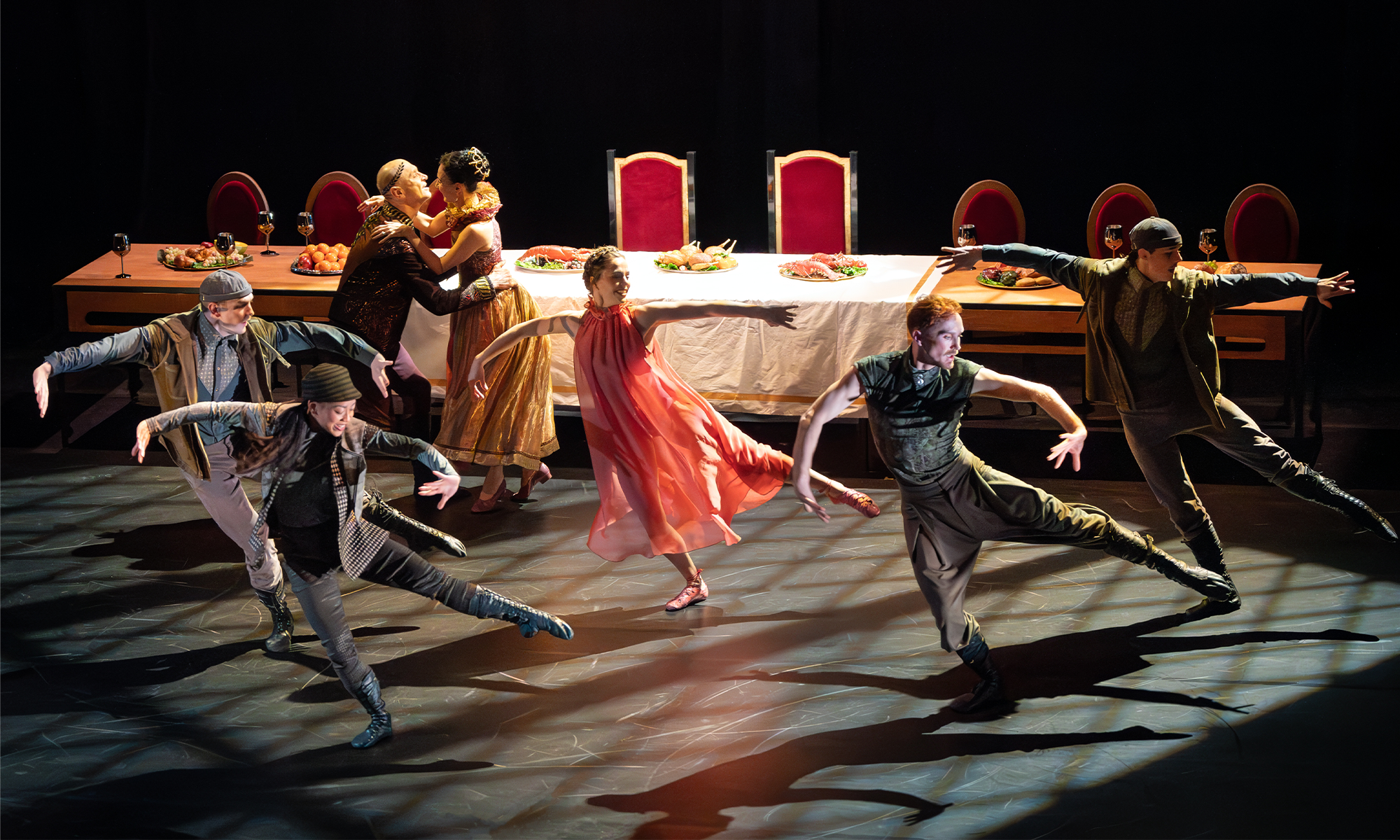 A group of dancers mid jump on stage. They stand in front of a long dining table.