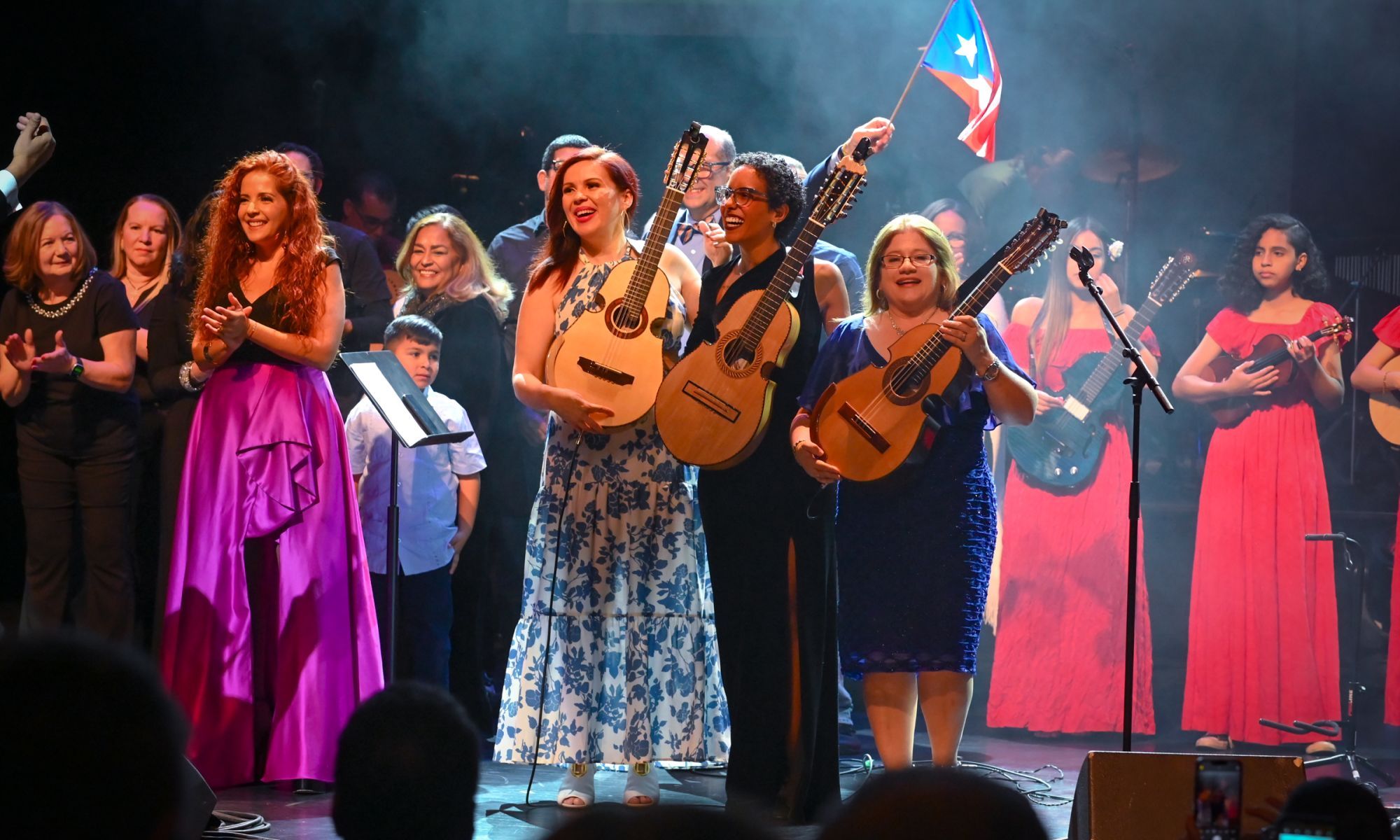 Cuatristas Maribel Delgado, Emma Colón Zayas, and Fabiola Méndez hold up their cuatros to the audience at the 25th Annual National Cuatro Festival as someone waves a Puerto Rican Flag in the back. 