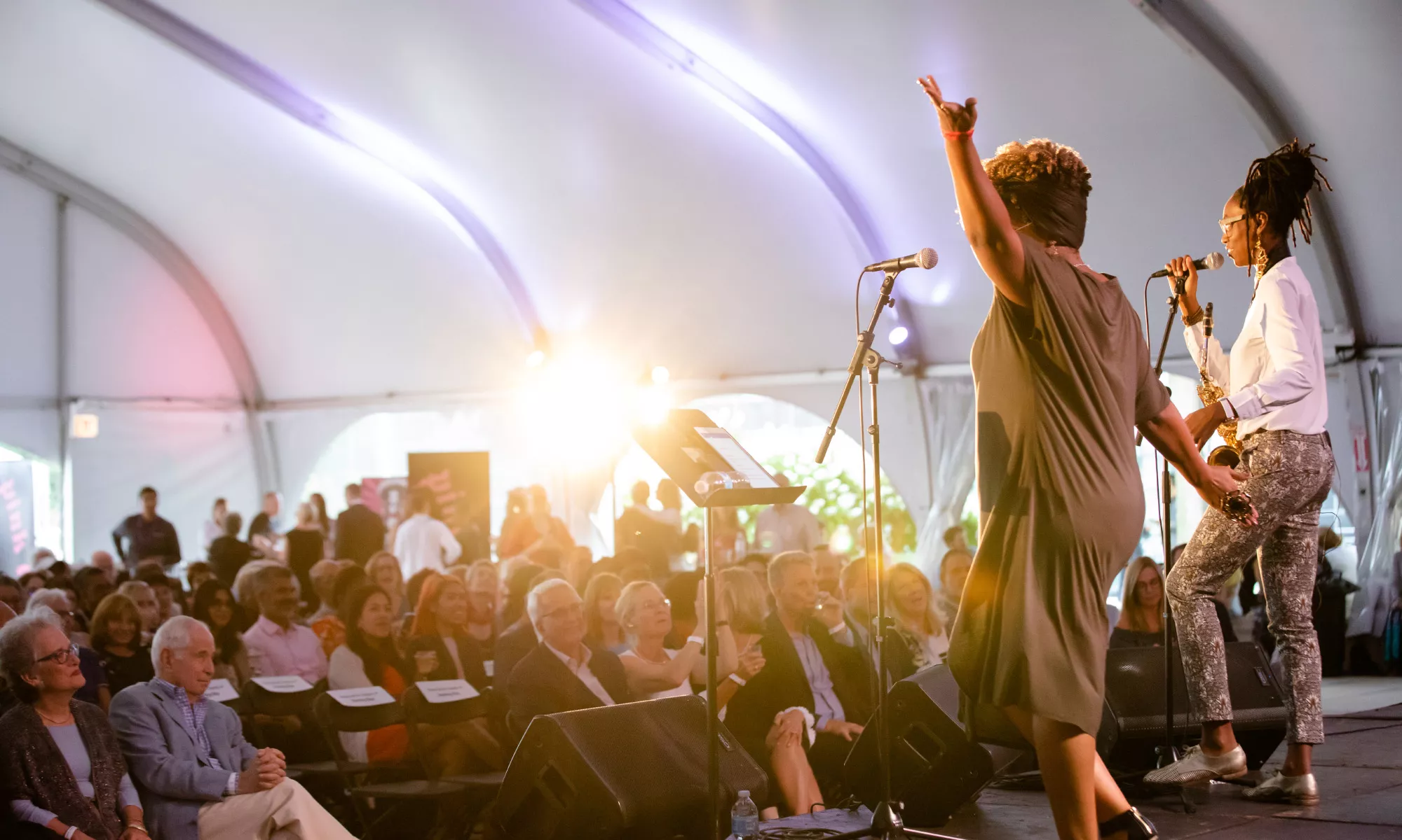 Two women sing on stage on the Harris Theater rooftop, performing to a room full of people.