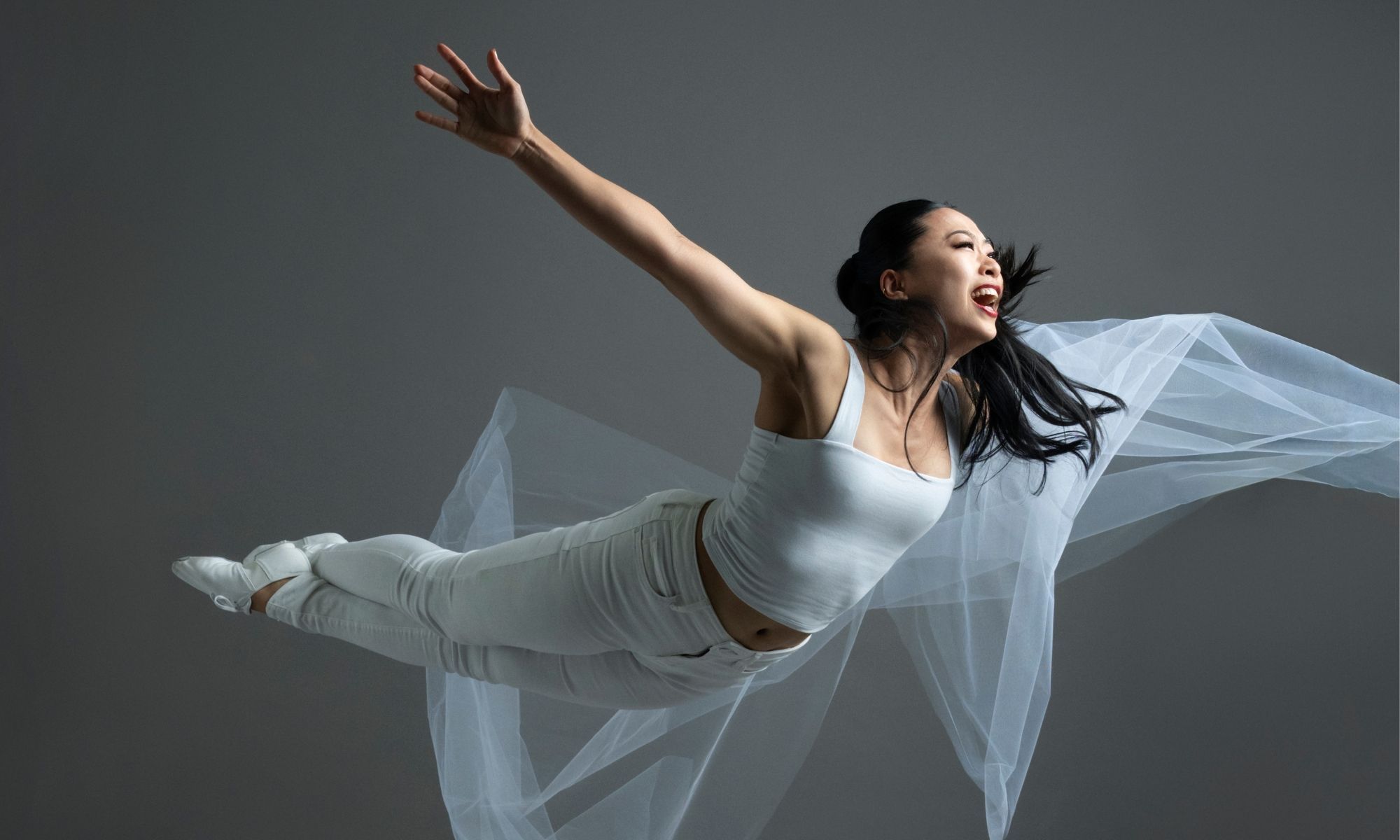 A dancer midair spread her hands open. She wears white pants and shit holding a white sheer piece of fabric. 