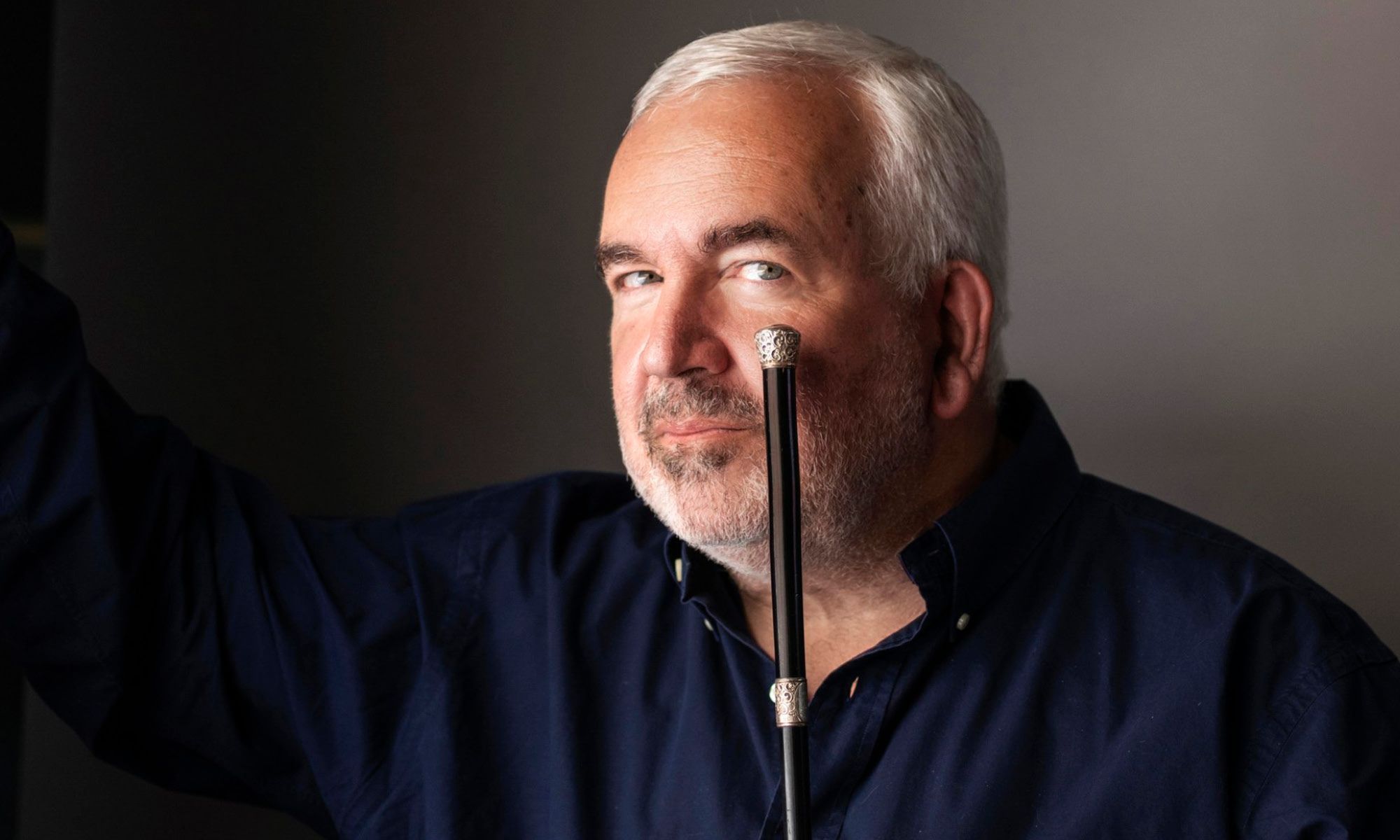 A gray-haired conductor holds a silver-tipped conducting baton to the corner of his mouth. 