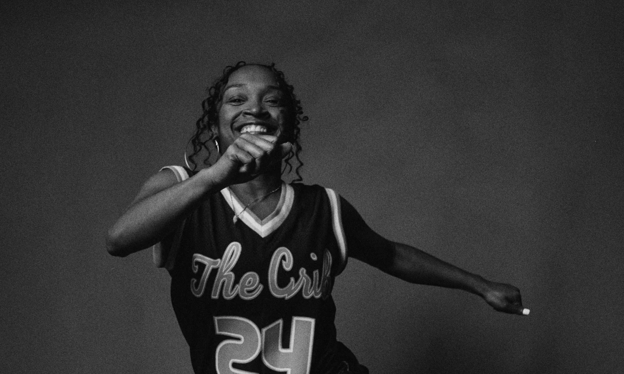 Black and white image of Donetta “Lilbit” Jackson dancing. Donetta smiles widely at the camera as she dances with her hands shuffling from the front to the side of her body. She wears a jersey, athletic shorts, and sneakers. The background is a deep grey making the darkness of her outfit stand out.