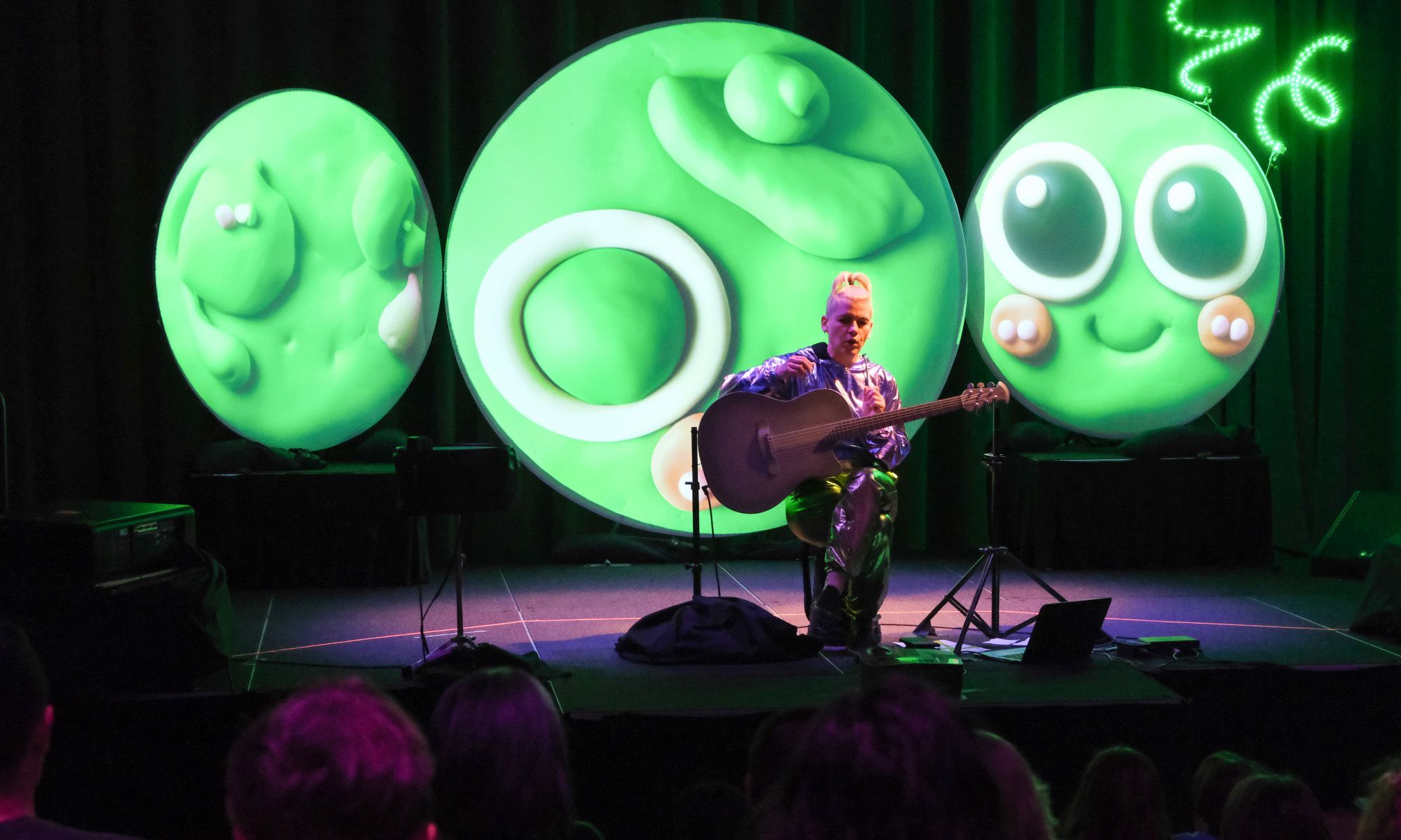 Kaki King holding a guitar performing in front of an audience. Three screens project a playful bug behind her.