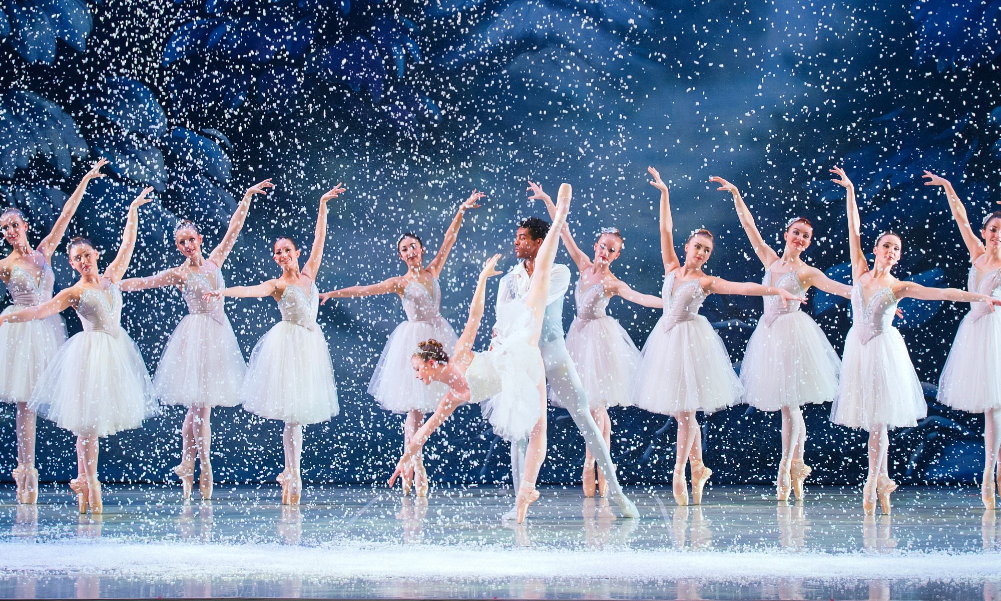 A vibrant scene from the Ballet Chicao's Nutcracker, showcasing dancers in elegant costumes amidst a magical winter backdrop. Snow is falling onstage as ballerinas pose on pointe. The Snow King and Queen are center stage.