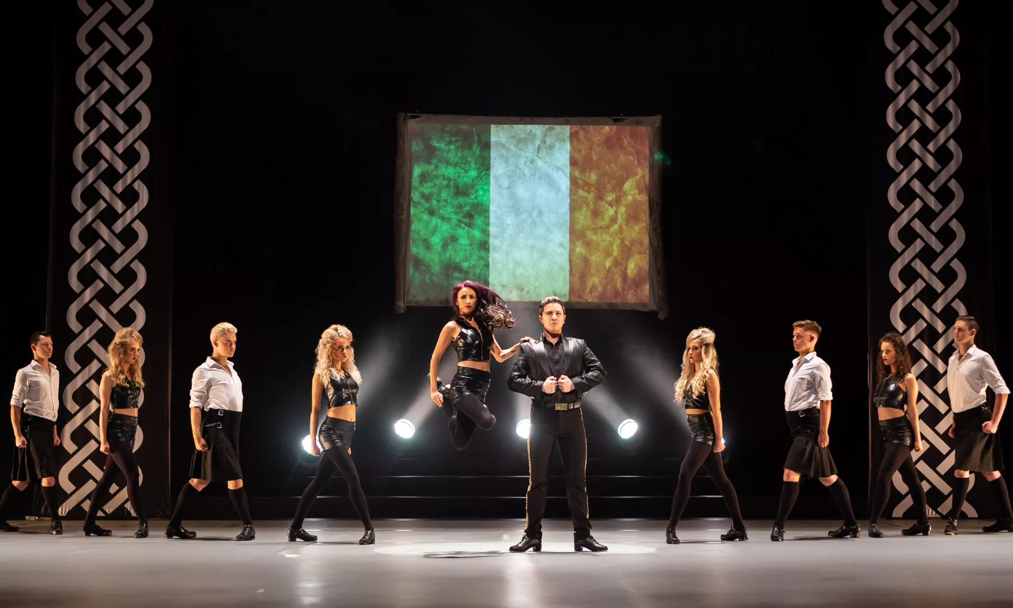  A dramatic stage performance featuring a line of dancers dressed in black and white costumes. A female dancer leaps into the air at center stage as the lead performers stand in pose. Behind them, a large Irish flag is projected, and two large Celtic knot banners frame the scene, reinforcing the show's Irish cultural theme.