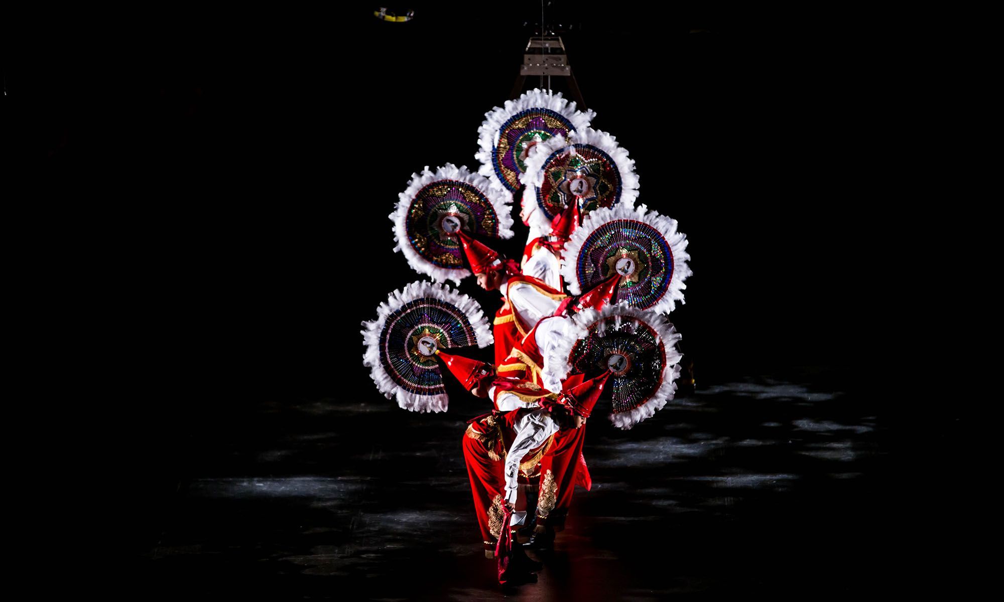 Dancers stand in a row wearing circular head pieces and bowing down.