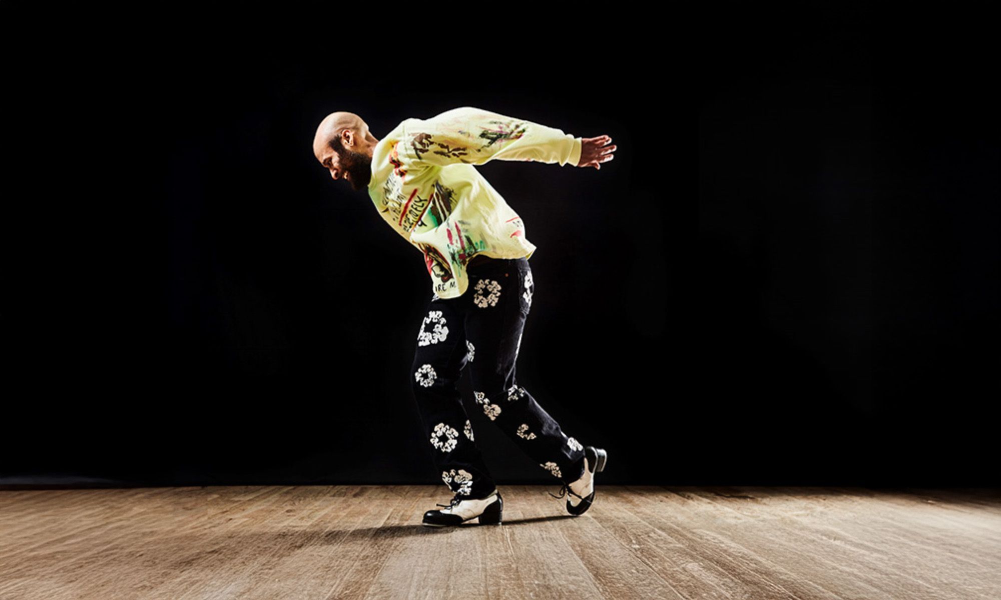 Jumaane Taylor dancing on a wooden floor. He wears black floral pants and a yellow printed long sleeve.