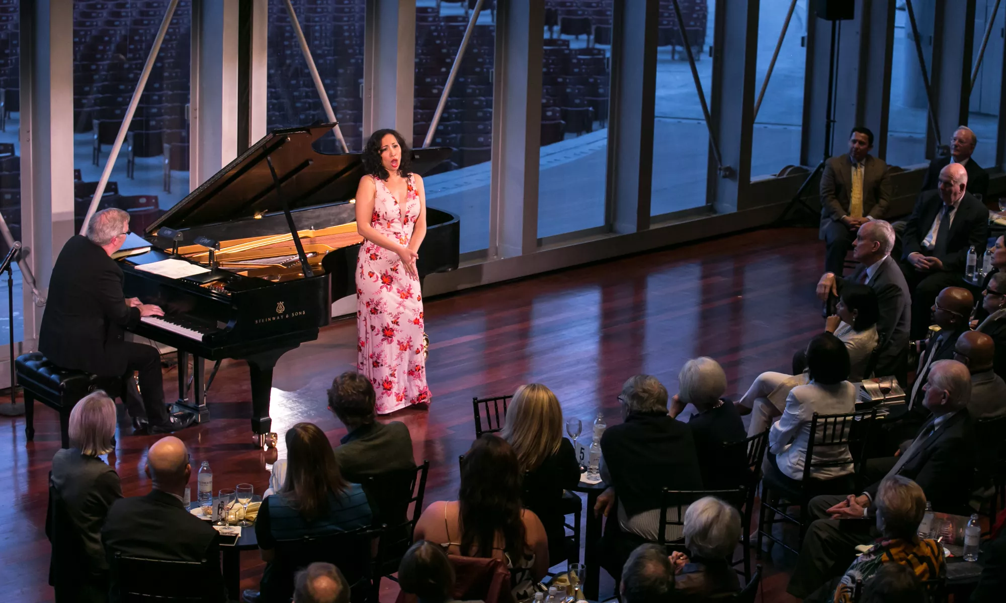 A woman wears a pink floral dress. She is singing in front of a seated audience and accompanied by a pianist.