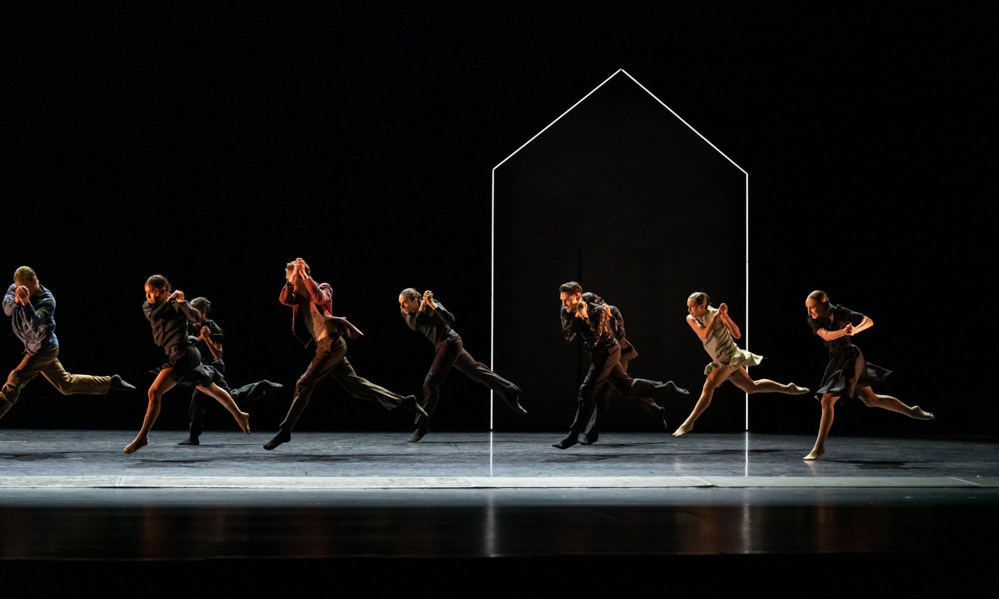 A group of dancer's mid performance jumping in the air. Only the dancers and an outline of a house is lit.