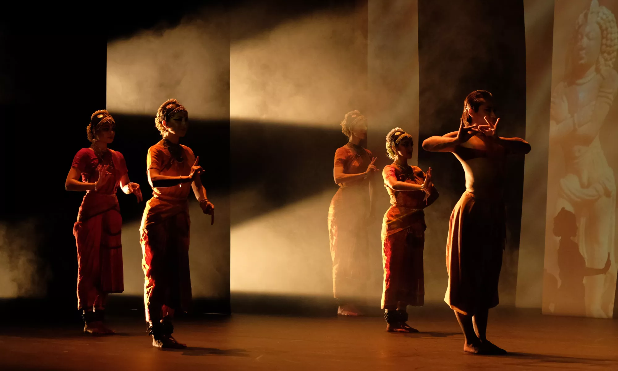 Five dancers stand wearing traditional Hindu attire. The stage is semi dark with the light coming through the back of the stage and large panels.