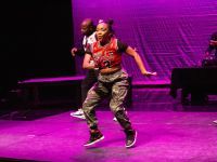 Image of Donetta “Lilbit” Jackson dancing on a brightly colored stage. Donetta appears mid movement with her hands shuffling from the front to the side of her body. She wears a Chicago Bulls’ jersey, a black undershirt, and camo-colored pants. The stage is lit a bright magenta, deepening the black shadows in the background. Other dancers are in the background and a dj attentively looks at his turntables.