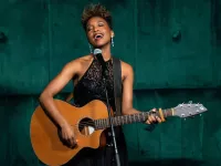 Image of Nashon singing. She appears mid-song with her mouth agape and is strumming a brown guitar. Nashon wears a long black dress and is photographed on a teal, velvet-like background.