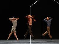 Three dancers on stage jump in unison while looking up and covering their ears.