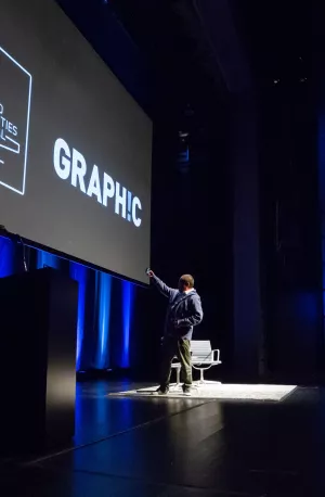 A man stands on a theater stage with blue accent lighting and a projected backdrop with white text that reads "Chicago Humanities Festival - GRAPHIC"