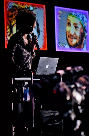 In the foreground, a woman stands in front of her DJ set up on stage, holding a microphone. In the background, colorful headshots of Lucky Plush dancers are projected, with borders of red, blue, purple. 